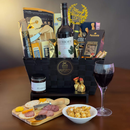 Gourmet gift basket with wine, snacks, and charcuterie board on a wooden surface.